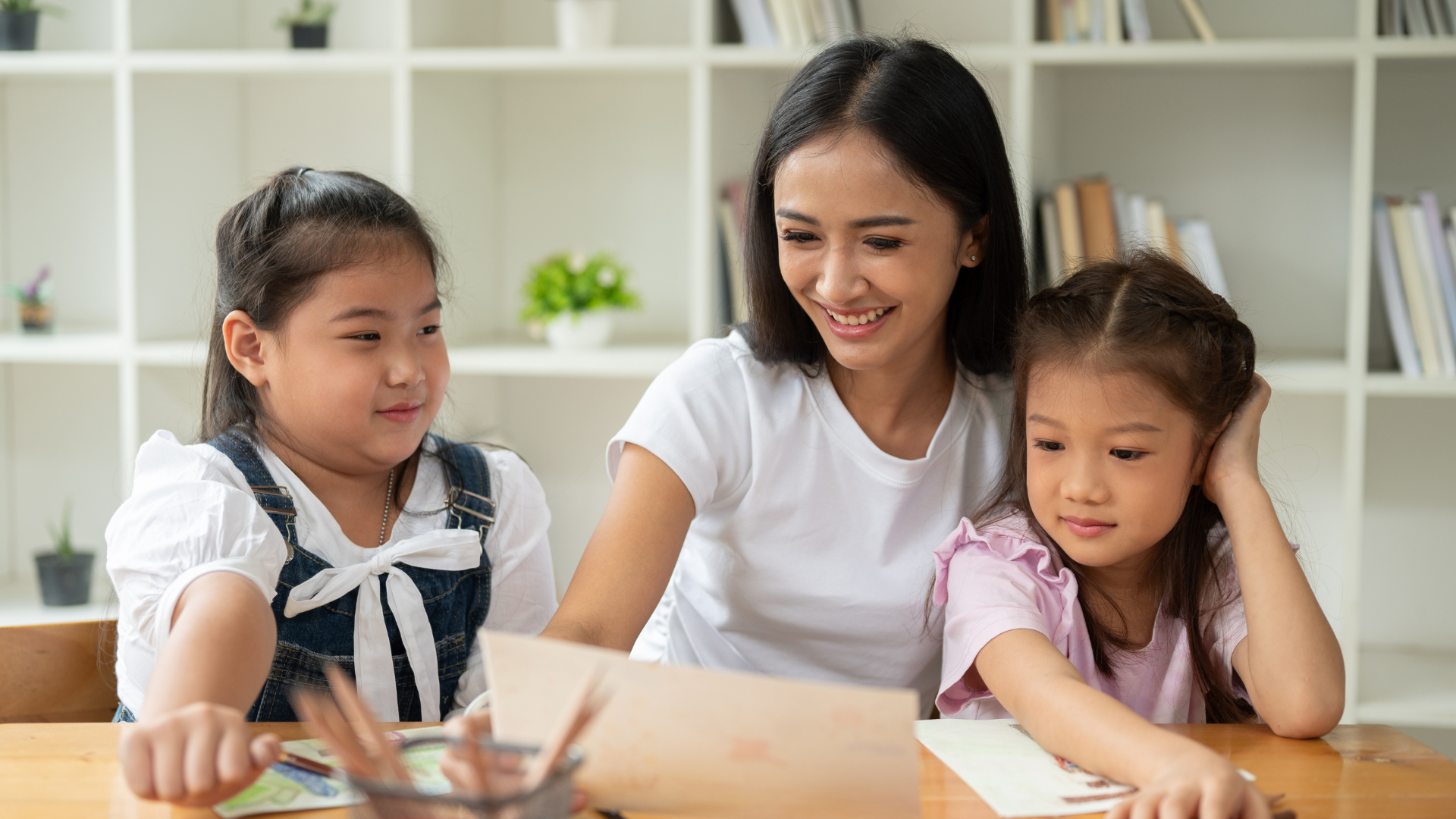 Seorang babysitter sedang mendampingi dua anak kecil saat belajar dan menggambar di meja.